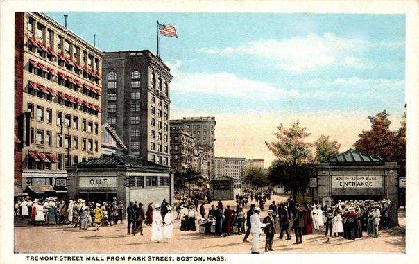 Vintage color postcard depicting Tremont Street Mall from Park Street in Boston, Massachusetts. The printed caption along the bottom reads “TREMONT STREET MALL FROM PARK STREET, BOSTON, MASS.”

The view looks south along a broad pedestrian mall from Park Street toward the downtown area. The scene is crowded with pedestrians, many dressed in early-20th-century clothing. Men wear dark suits, long coats, and hats, while many women wear long dresses, coats, and elaborate hats. Several people carry parcels and luggage, and groups of pedestrians are gathered near the entrances to the underground transit station.

On the left is a large multi-story masonry commercial building with rows of windows fitted with red-and-white striped awnings. At street level are small storefronts and a prominent stone-and-glass entrance structure marked “OUT.” Directly behind it is a taller dark-brick building with large arched windows near its upper floors. An American flag flies from a pole above the building.

On the right is a matching stone entrance structure prominently marked “SOUTH BOUND CARS ENTRANCE.” This structure has a low, hipped roof with a series of skylights and appears to provide access to the underground streetcar or subway system. Large crowds surround both the entrance and exit structures.

The center of the image opens onto a landscaped pedestrian area with trees, benches, and additional small transit structures. More pedestrians fill the mall in the distance, with several large downtown buildings visible beyond the trees. The architecture, clothing, streetcar entrances, and crowds give the scene the appearance of Boston's early public-transit era, likely around the first decades of the twentieth century.

The postcard is especially notable for documenting the above-ground architectural treatment of Boston's early subway entrances at Park Street, integrated into a busy pedestrian environment surrounded by downtown commercial buildings.
