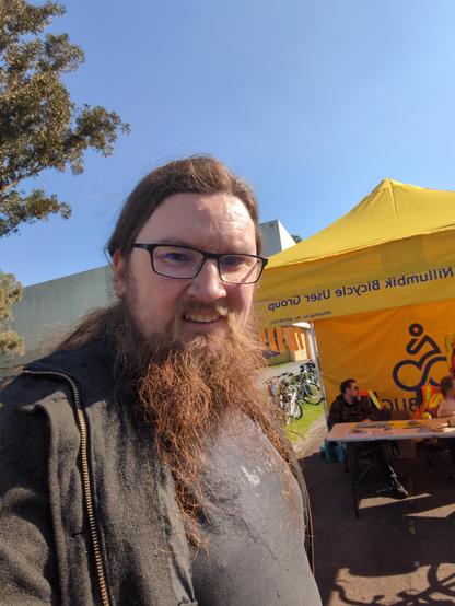 A selfie of a man with a beard and dark rim glasses (me) standing out side the bicycle valet parking. The marquee is yellow and has "Nillumbik bicycle user group" written on it.