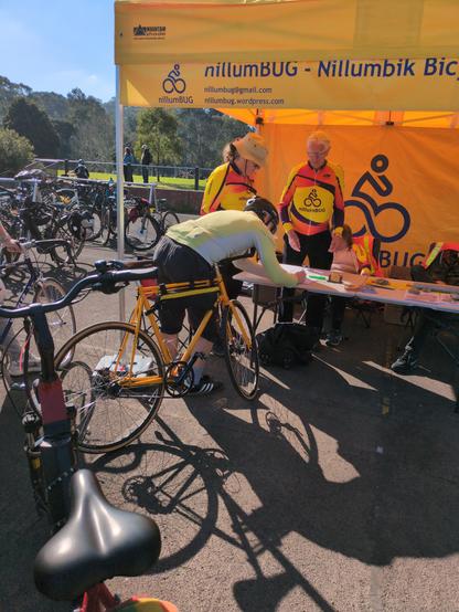A photo of the valet parking marquee. A man with a yellow bicycle is signing in. In the background there are racks of bicycles.

The people running the marquee are wearing yellow shirts with the Nillumbik bicycle users group logo.