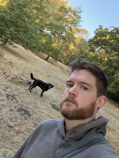 A selfie of a man wearing a grey hoodie. He has a dirty blue dog lead over one shoulder. His ear is pierced. 

In the background looking at the camera is a charcoal Labrador. The ground is dry and dead. There are trees and morning sun.