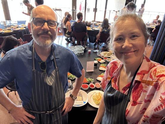 Two middle aged people in aprons in front of a table with pre-prepared kimchi ingredients