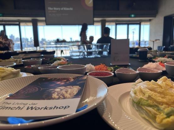 A table in the foreground has plates with salted cabbage and a recipe book that says: The Living Jar - Kimchi Workshop. Lots of bowls are visible. The background looks out windows into a sunny harbour scene.