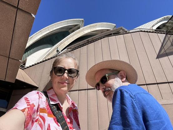 A man in a blue shirt and straw hat next a woman in a pink shirt, standing in bright sun under the sails of the Sydney Opera House