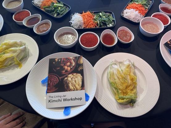 Looking down at a table with plates with salted cabbage and a recipe book that says: The Living Jar - Kimchi Workshop. Four bowls hold spices and pre-mixed sauces. A platter holdes grated vegetables.