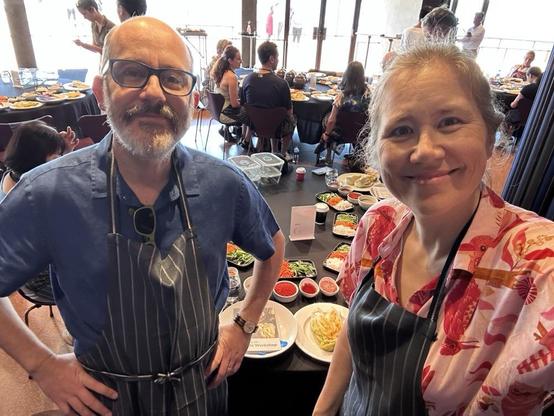 A man in a blue shirt next a woman in a pink shirt, with a table of food behind them. Both are wearing blue aprons.