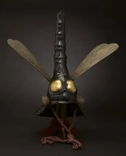 The helmet features a tall, ridged central crest that forms the body of a dragonfly, flanked by wide wooden wings. Two large, rounded eyes finished in gold leaf sit prominently on the front.