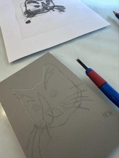 A piece of grey lino with a drawing of a cat on it, next to a carving tool