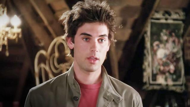drew fuller as chris on charmed. he’s wearing a khaki jacket over a red t-shirt and is standing in the attic of halliwell manor