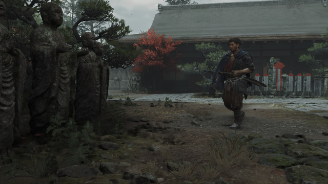 Musashi walking past stone Jizo statues near a traditional temple with autumn foliage.