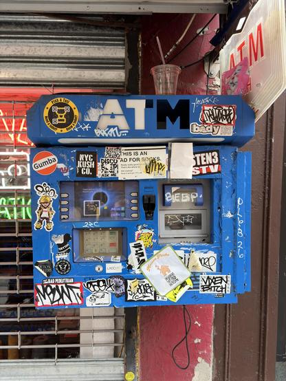 Blue ATM kiosk covered in stickers and graffiti, with a clear plastic cup and straw on top, located outside a building.