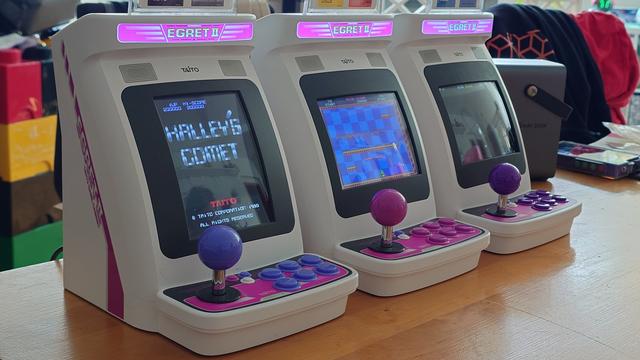 three mini arcades on a table.
all of the same Taito make, but the sticks and buttons are in different shades ofnpur0le, blue and pink.