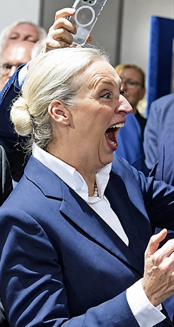 A photo of a blonde woman in a blue suit and white shirt celebrating with an open mouth