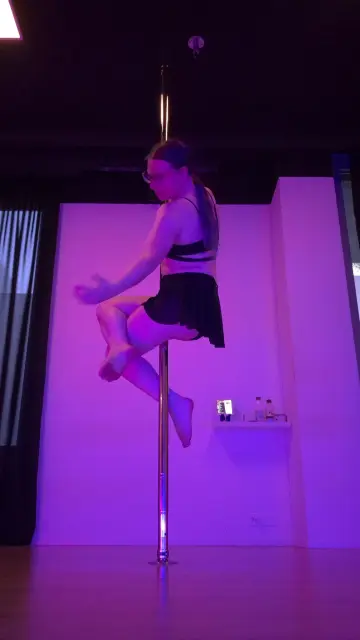 Alicia sitting halfway up on a pole, stretching one arm out while her legs are crossed.