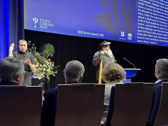 A First Nations man addresses an audience from a lectern. An AUSLAN interpreter stands beside him.