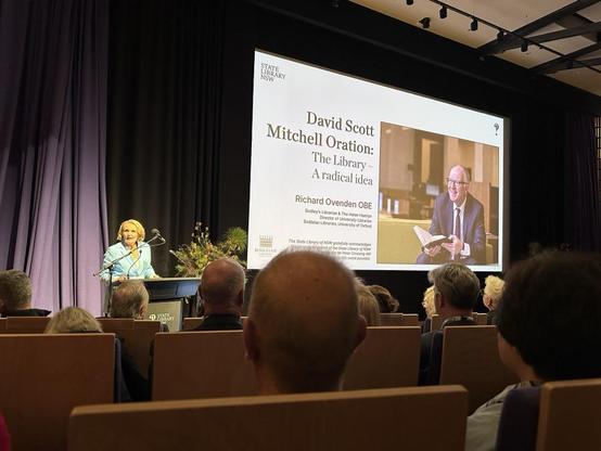A woman in a blue coat behind a lectern in an auditorium. The slide reads: David Scott Mitchell Oration: The Library - A radical idea Richard Ovenden OBE