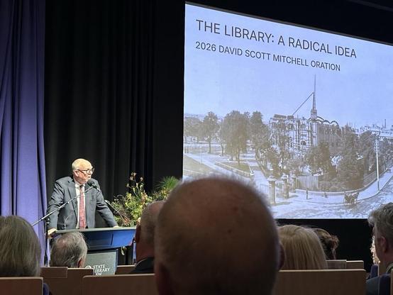 A man presenting at a lectern. The slide behind him reads: The Library: A Radical Idea 2026 David Scott Mitchell Oration