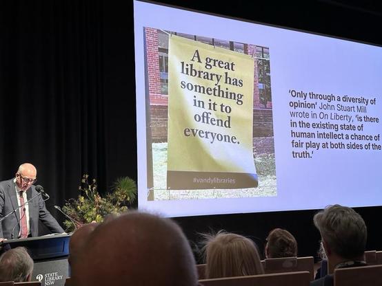A man presenting at a lectern. The slide behind him shows a banner that reads: "A great library has something in it to offend everyone" as well as a quote: 'Only through a diversity of opinion' John Stuart Mill wrote in On Liberty, 'is there in the existing state of human intellect a chance of fair play at both sides of the truth.'