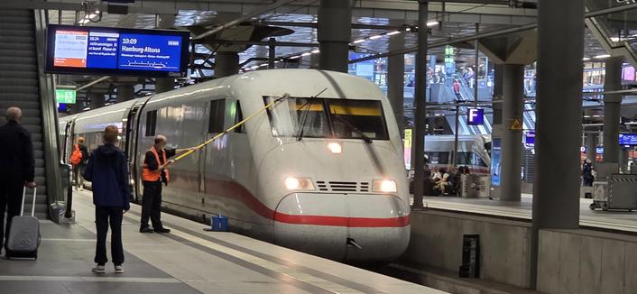 Motiv: Ein ICE 1 steht an einem überdachten Bahnsteig (Gleis 7) mit Zielanzeige „Hamburg-Altona“.
Details: Ein Bahnmitarbeiter in Warnweste reinigt die Frontscheibe des Triebkopfs mit einer langen Teleskop-Wischstange. Auf dem Bahnsteig stehen Reisende mit Koffern.