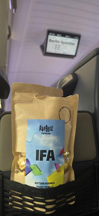 Eine geöffnete braune Papiertüte Popcorn der Berliner Marke „Knalle Popkorn“ steckt im Sitztaschennetz eines Zuges.

​Details: Das blaue Etikett trägt den Aufdruck „IFA“ sowie die Sorte „Butterkaramell Tahiti-Vanille“. Im unscharfen Hintergrund ist ein Deckenbildschirm mit der Haltestellenanzeige „Berlin-Spandau“ zu sehen.