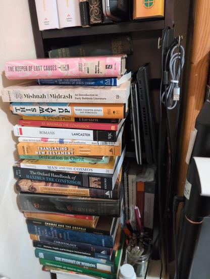 Photo of stack of books next to bed about 3 feet 1 meter high in front of a bookcase and next to another, with lamps in the foreground.