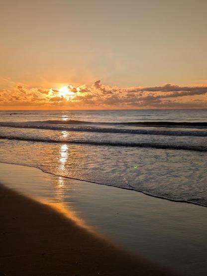 Photo of the sun rising over a calm ocean at the edge of the beach. The sun is bright orange, hidden behind a ribbon of clouds. Its reflection is mirrored on a line of gentle waves. In the foreground is a thin strip of wet sand, where the surf washes over and recedes.