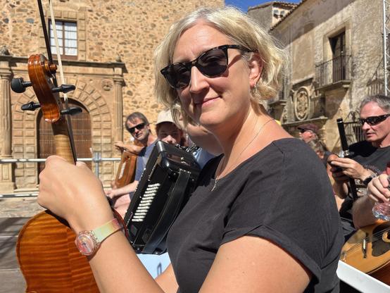 Jessica in full sunlight holding her fiddle sitting on stage with rows of other musicians.