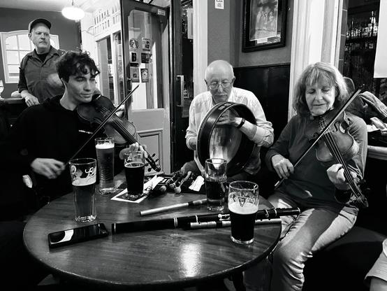 A bodhrán player between two fiddlers at a pub table with pints and a flute on it.