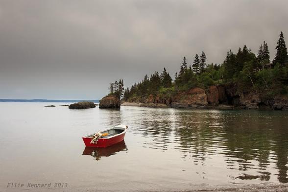 A single small red boat sits empty on the still waters of a bay at full tide. Its outboard motor is tipped up. There is a single paddle almost hidden behind the motor. 
Beyond the boat are a few rocks jutting up from the water, with some more so, and a single one tall enough that there are a couple of scrubby pines growing on top of it. To the near right of the frame is a wooded shore reflected in the water, and in the far distance a low shore is just visible through a light mist. The sky is cloudy grey and the water has the same dull tone, broken only by the bright red of the boat. It is a peaceful scene.