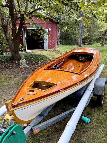 A white hulled handmade wooden boat with gleaming varnished deck, trim, and interior, on a trailer in a yard with an old shed in the background.