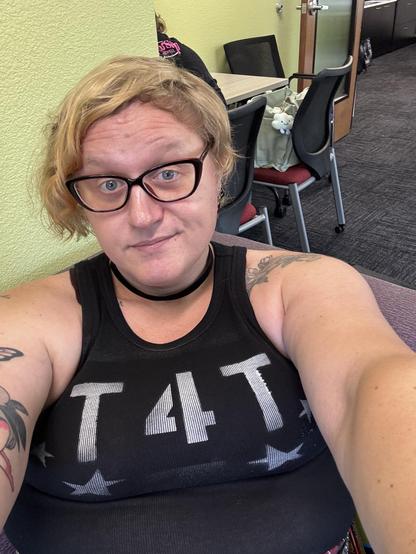 Selfie of me in a black tank top that has t4t printed in white letters, as well as a black choker and dark red glasses with a bit of a cateye style. My blond hair is asymmetrical and a bit naturally disheveled and curling about in different angles