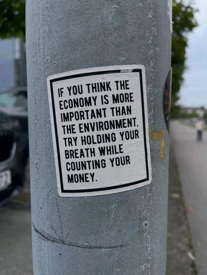 A white sticker on a grey streetlamp reads: “If you think the economy is more important than the environment, try holding your breath while counting your money.