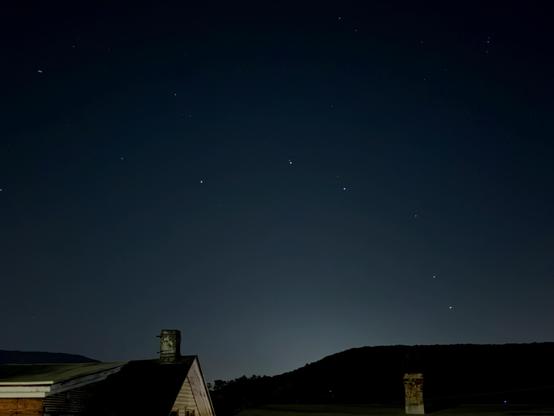 Starry night sky above a roof with a chimney and dark hills.