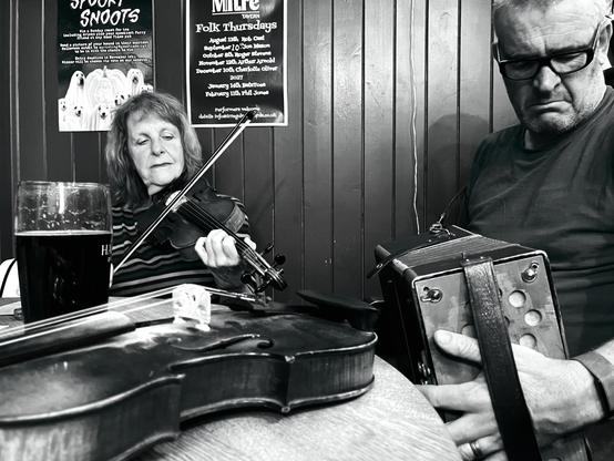 A man playing button accordion and a woman playing fiddle at a pub table with a fiddle on it.