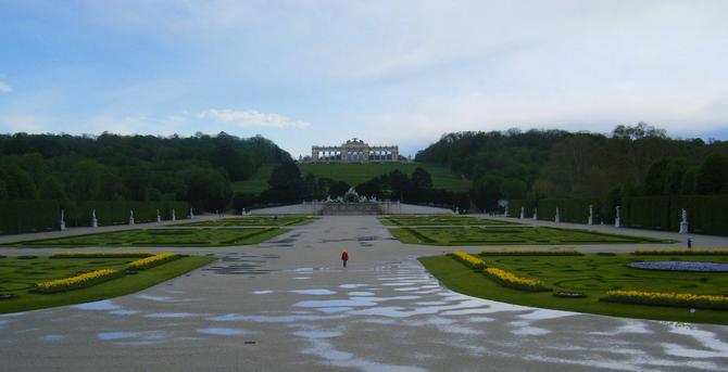 Wide view over a deserted, rainy park full of monuments. There is one person in the distance, walking away from the camera, wearing a red jacket. This was taken in the Spring, so there are some flower beds in blossom.