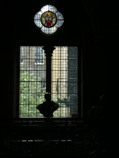 Photo taken from inside a dark stairwell of a view out of a window of many small panes of glass. There is a bust in silhouette in front of the window. Above the rectangle of the main window there is a kind of "bullseye" window, shaped like a flower with four petals. At the centre of this is some stained glass, featuring Scotland's flag. Outside you can see a low tree and the rear of a stone building with traditional Edinburgh windows.