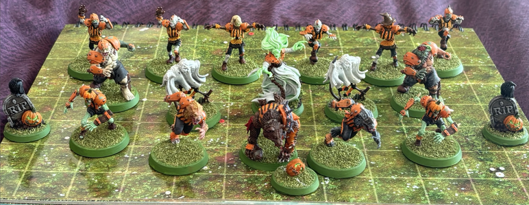 A team of miniatures with green grass bases, stood on a grassy board. There are 16 miniatures; zombies, werewolves, ghosts, ghouls a banshee. All are in an orange and black team strip.