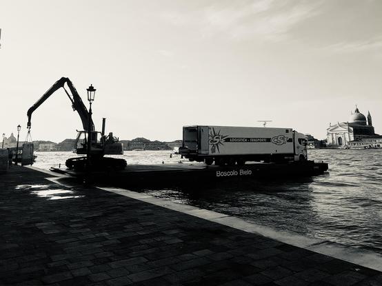 Black and white photo of a long floating platform in a wide canal, berthed by a stone pavement. On the far end of the platform is a big white lorry, and on the pavement end, a small crane is busy winching goods onto shore. In the background on the far side of the canal is the imposing bulk of the Redentore church, providing a stark ancient-and-modern and sacred-and-profane contrast.
