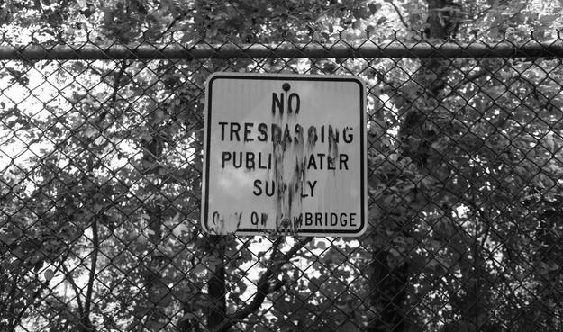"No Trespassing, Public Water Supply, City of Cambridge"

The road sign's text is smudged a bit, as if water had melted it over time. The sign is on chain-link fencing, behind which are trees and shrubs.