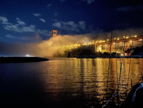Golden Gate Bridge lit by streetlights with fog wafting in on the breeze. A ship is passing out the gate from left to right. Multiple fog horns are sounding. Lights reflected on dark water and light puffy clouds against a dark sky. The phone took a while to gather enough light.