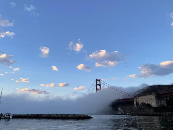 The north pier of the a Golden Gate Bridge pokes up into blue sky above a dark fog bank pouring in the Gate. A stone breakwater separates the calm water in the foreground from the choppy water stirred up by the fog wind. A few white puffy clouds dot the sky.