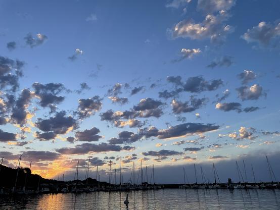 Orange light beams flash up from lower left of photo, illuminating numerous small puffy clouds that are mostly dark. Sailboat masts along the bottom are silhouetted against a dark fog tongue being blown in the Golden Gate. The masts are also reflected in the water in the foreground.