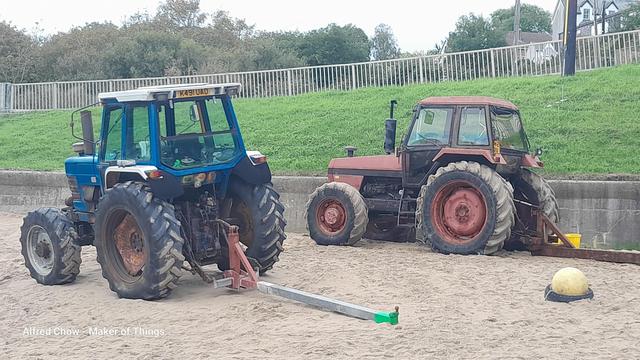 Two big old four wheel drive tractors parked on the beach by a concrete wall. A blue one on the left and a red one on the right, both facing left and seen from 3/4 rear.
They are rusty and set up for boat launching.
The background shows a grass embankment rising up to a metal fence to the road, with trees beyond.