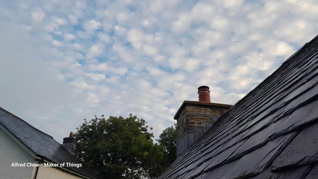 A view over a slate roof top, on the right, with a stone chimney at the end of the ridge.
On the left is the edge of the slate roof on a cream coloured house.
In between is the crown of a bushy green tree.
The sky above is blue with a covering of cotton wool ball clouds.