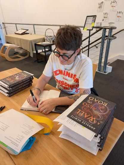 It is I, smiling, as I sign pre-order copies of my book upstairs at Green Apple Books. I am wearing a t-shirt and sitting at a pine wood desk.