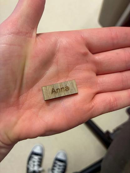 Anna’s hand holding a tiny engraved rectangle of birch that reads “Anna.” Her black converse shoes are on the floor out of focus in the shot too