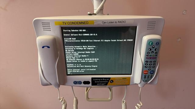 A photograph of a hospital TV with two phone-like attachments on either side. Everything is beige. There is a yellow sticker on the top that reads TV CONDEMNED. The screen is black with white text showing DOS errors.