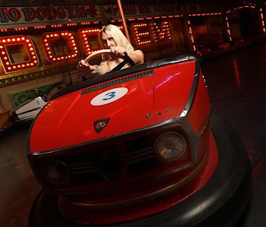 A model in a dodgem car in front of an illuminated fairground sign "Dodgem".  The angle is a little dramatic.