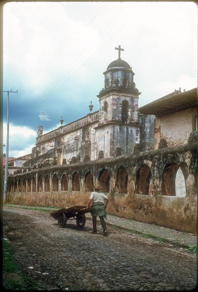 A man pushes a wheelbarrow along a cobblestone road next to an ancient church. An ominous dark cloud is on the horizon/