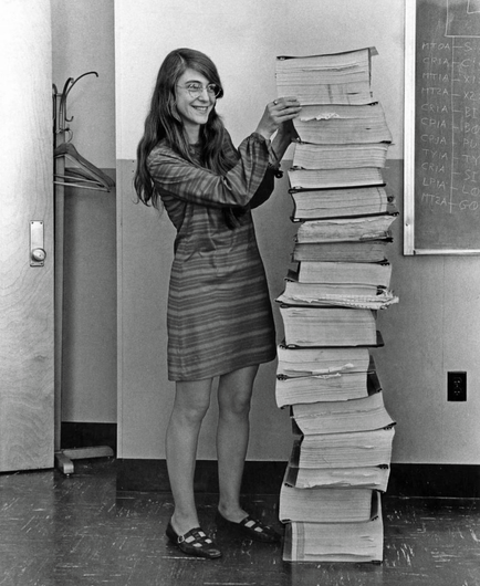 A black and white photo picture from the 60s of a woman stacking piles of printed out computer code.  This woman is Margaret Hamilton.