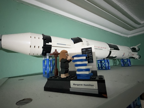 A photo of my Lego SaturnV rocket on a dusty shelf.  In front of it is a Lego version of the photo.  Margaret Hamilton stacking piles of paper.